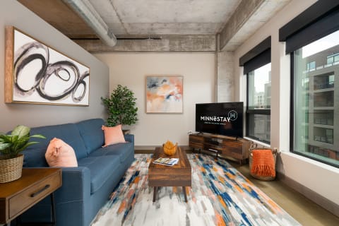 A stylish living room featuring a blue sofa, colorful artwork, and a TV displaying Minnestay logo.
