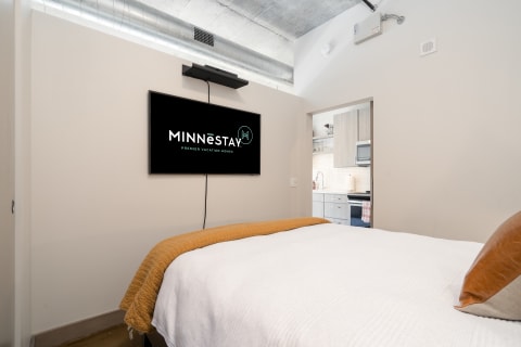 Modern bedroom with a double bed, orange throw blanket, and a flat-screen TV displaying the MINNēSTAY logo.