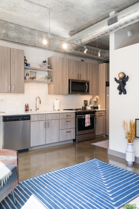 A modern kitchen with light wooden cabinets, stainless steel appliances, and a vibrant blue striped rug.
