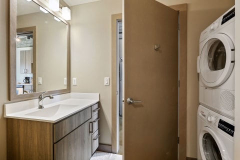 A modern bathroom with a wooden vanity and laundry machines in the background.