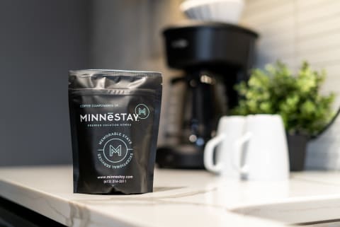 Black coffee bag from MINNēSTAY with coffee maker and white cups in the background.