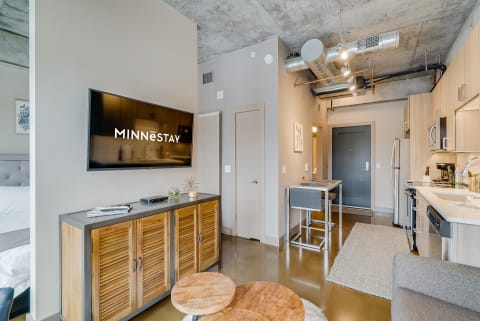 Modern studio apartment with a TV, wooden cabinet, dining area, and kitchen appliances.