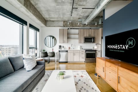 Modern living room with a gray sofa, kitchen space, and a large TV displaying MINNESTAY logo.