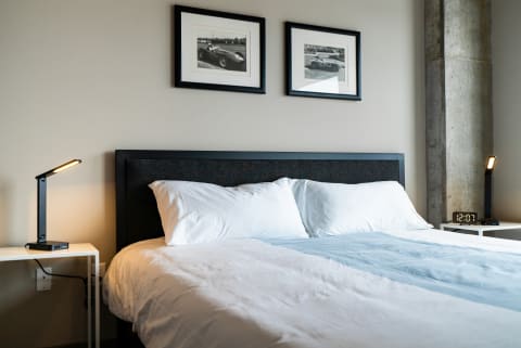 A neatly made bed with white pillows and a blue bedspread, framed photos on the wall, and a lamp on a nightstand.