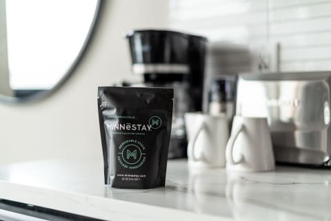 A black coffee pouch from MINNESTAY stands on a marble countertop with white mugs in the background.