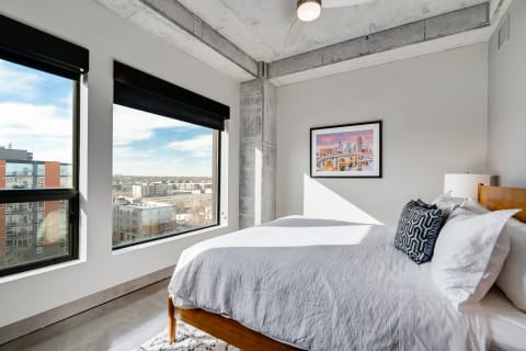 Modern bedroom with large windows, a neatly made bed, and cityscape artwork.