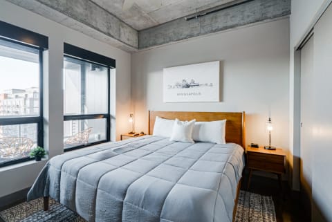 A stylish bedroom with a gray bedspread, wooden furniture, large windows, and a decorative Minneapolis artwork.