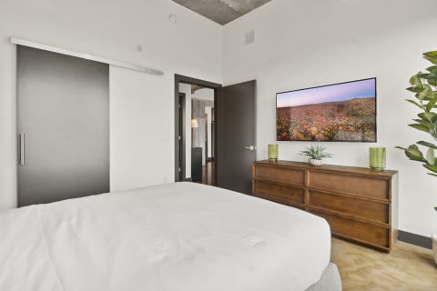 A modern bedroom featuring a white bedspread, wooden dresser with green vases, and a wall-mounted TV displaying a landscape.
