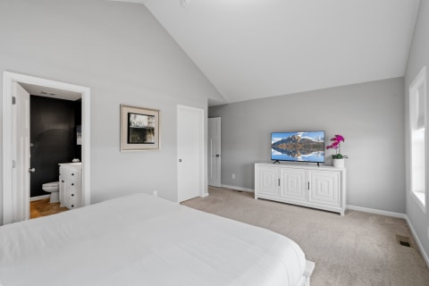 A spacious bedroom with a white bed, modern TV, and a purple orchid, providing a tranquil atmosphere.