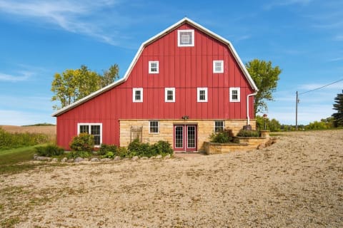 Red barn with stone base and green foliage in a rural setting under a blue sky.