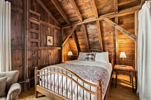 A cozy bedroom with wooden walls, a sloped ceiling, a decorative bedspread, and a soft armchair.