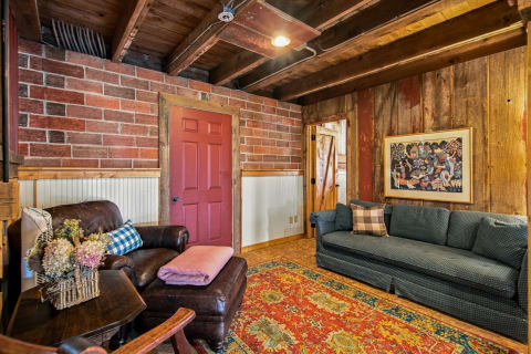 A cozy living room featuring a rustic design with exposed beams, a maroon door, a leather armchair, and a vibrant rug.