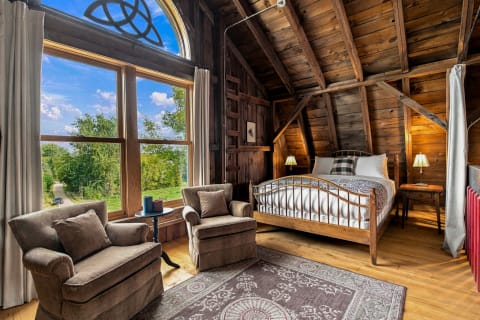 A rustic bedroom featuring a large window with countryside views, wooden ceiling, and cozy furnishings, perfect for relaxation.