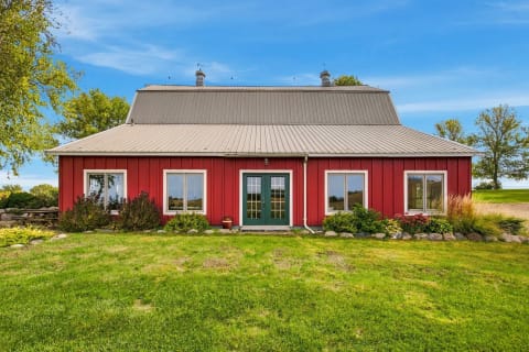 A red barn-style house with a gray metal roof, surrounded by green lawns and colorful flower beds under a clear blue sky.