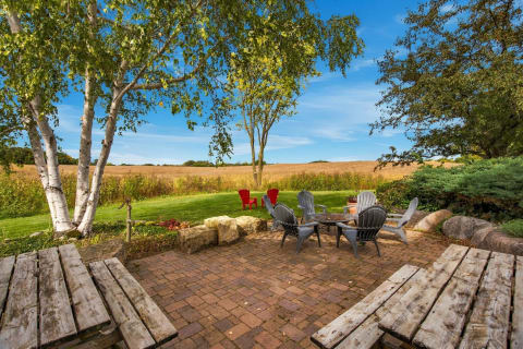 A cozy outdoor patio with grey Adirondack chairs, a fire pit, and birch trees overlooking open fields.