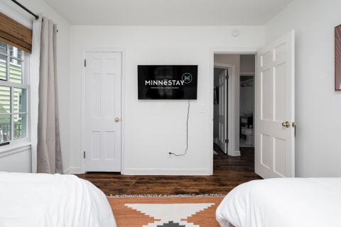 A contemporary bedroom featuring two beds, a flat-screen TV displaying the MINNESTAY logo, and warm wooden flooring.