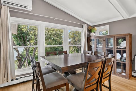 A cozy dining room featuring a polished wooden table, wooden chairs, and large windows with a garden view.
