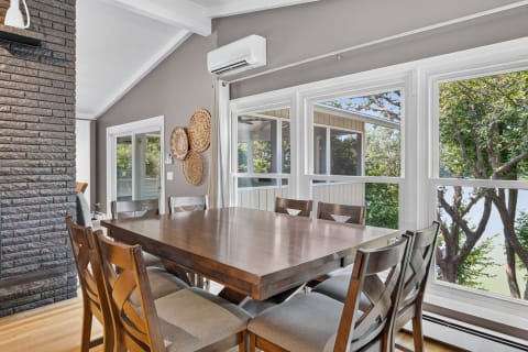 Spacious dining area with a wooden table, chairs, and large windows showing lush greenery outside.