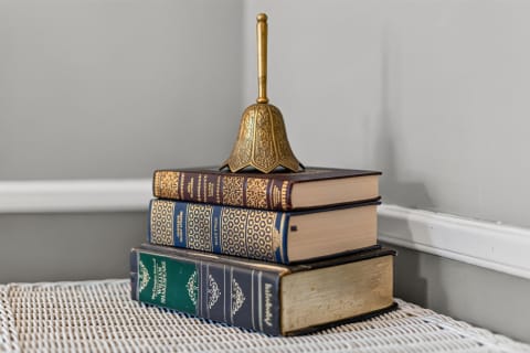 A stack of classic hardcover books with a brass bell on top in a cozy room corner.