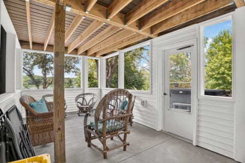 A cozy screened porch featuring wicker chairs with teal cushions, wooden beams, and large windows overlooking greenery and a lake.
