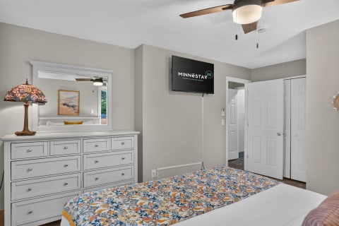 A cozy, well-decorated bedroom with a colorful comforter, white dresser, ceiling fan, and mounted TV.