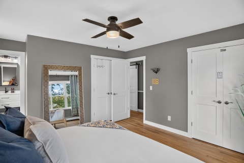 An elegant bedroom with gray walls, wooden floors, a ceiling fan, and a large mirror reflecting a window.