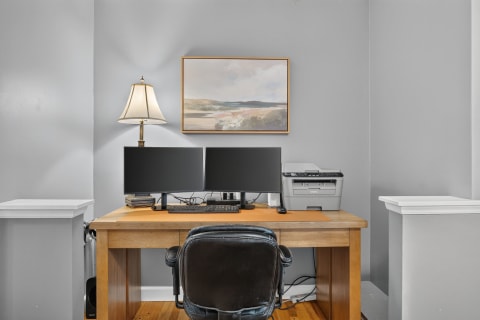 A wooden desk with two monitors and a lamp in a gray-walled workspace.