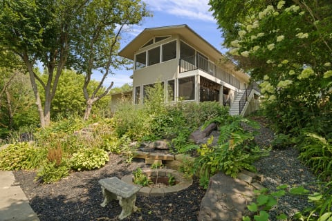 A two-story house nestled among trees with vibrant plants and a stone pathway.