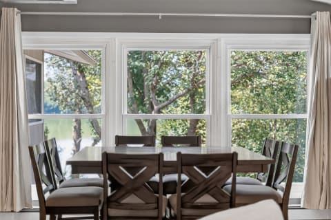 A cozy dining room with a wooden table, surrounded by X-back chairs and large windows displaying green trees outside.