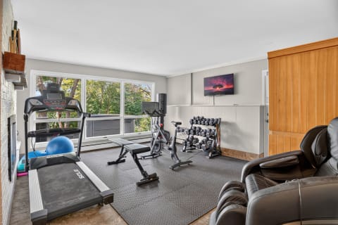 A private workout space with exercise equipment including a treadmill, stationary bike, dumbbell rack, and a massage chair, illuminated by natural light.