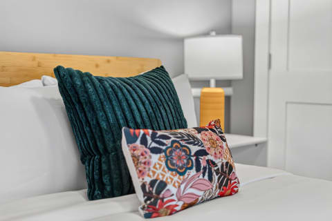 A stylish bedroom featuring textured teal and floral pillows on a white bed, with a warm lamp in the background.
