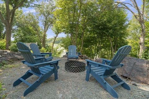 Three blue Adirondack chairs arranged around a fire pit with a lake view surrounded by greenery.