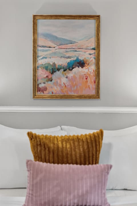 A cozy bedroom with white linens, decorative pillows, and a colorful landscape painting on the wall.