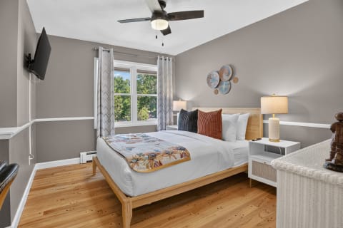 A cozy bedroom with a bed, lamp, ceiling fan, and decorative accents, illuminated by natural light.