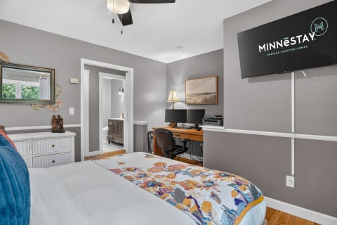 A cozy bedroom featuring a floral-patterned bedspread, workspace with dual monitors, and a flat-screen TV.