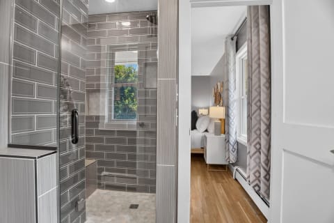 Walk-in shower with gray tiles and natural light, leading to a cozy bedroom.