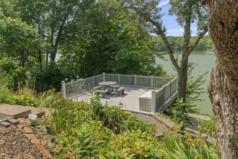 A peaceful lakefront deck with wooden furniture, surrounded by trees and greenery, overlooking a calm lake.
