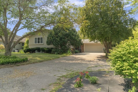 A suburban house with a green lawn, trees, and a driveway leading to a garage.