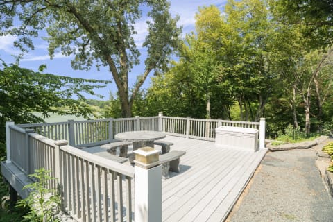 A spacious wooden deck with a stone table and benches, surrounded by green trees and a gentle pathway leading away.