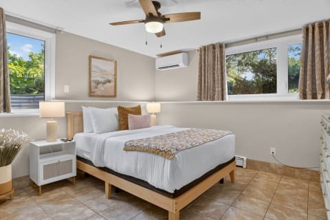 A restful bedroom with white linens, warm wood elements, and natural light from two windows.