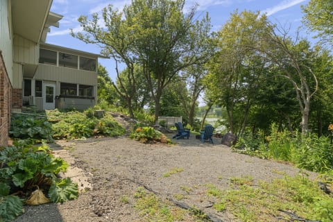 Gravel patio with two blue chairs surrounded by greenery and trees, overlooking a body of water.