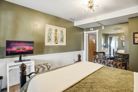 A cozy bedroom with an olive green wall, an iron bed, a flat-screen TV, and a stained-glass window.
