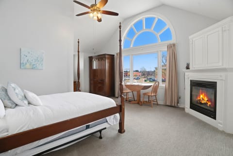 A spacious bedroom with a four-poster bed, a wooden wardrobe, and a fireplace, illuminated by large windows with a view outside.