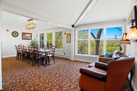 Dining area with a large table, wooden chairs, and seating arranged to view lush hills through large windows.