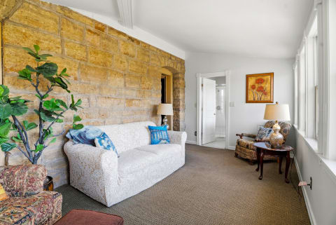 Comfortable seating area with a white sofa, patterned armchair, and stone walls, adorned with artwork and a potted plant.