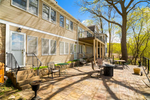 A spacious brick patio with seating and a two-story home amidst trees and blue sky.