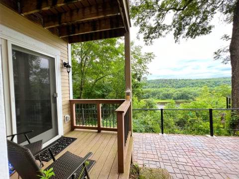 A private deck with wicker chairs overlooking green hills and a river beneath a cloudy sky.