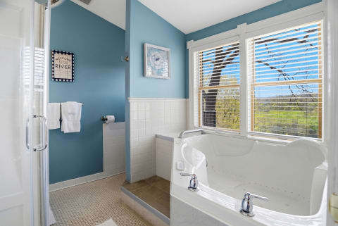 A serene bathroom featuring a jetted soaking tub, a shower, and large windows with views of the countryside, decorated in soothing blue and white hues.