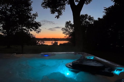 A hot tub with blue lights and a view of a sunset over a lake with silhouetted people on a dock.