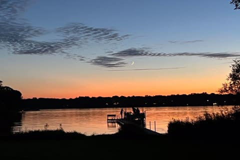 Silhouetted figures on a dock at twilight, with a crescent moon in the sky and colorful reflections on the lake.